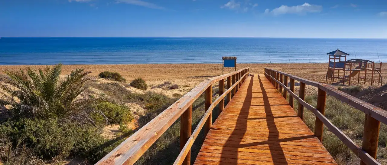 Pasarela de madera hacia la playa del rebollo, con arena dorada, mar azul y un pequeño parque infantil.