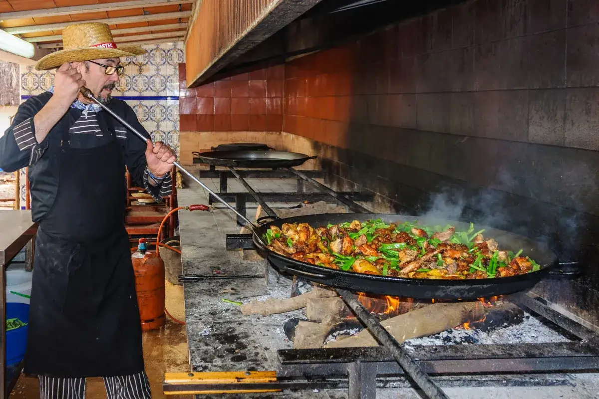 Chef con sombrero de paja prepara la mejor paella en Valencia, cocinada a leña en una gran paellera humeante.
