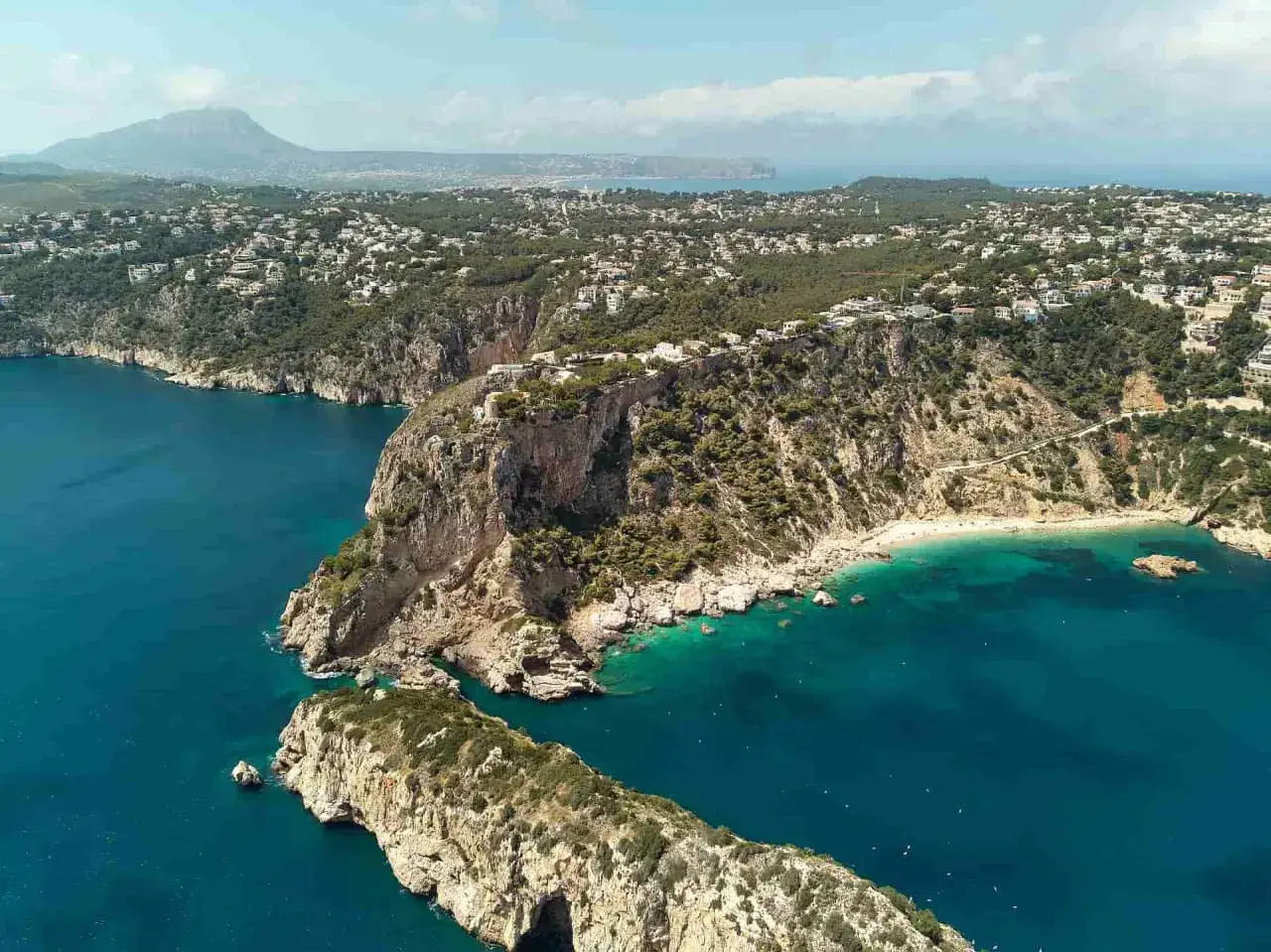 El mar de Jávea se extiende azul turquesa, abrazando acantilados rocosos cubiertos de vegetación y casas blancas. Una cala escondida invita al descanso.