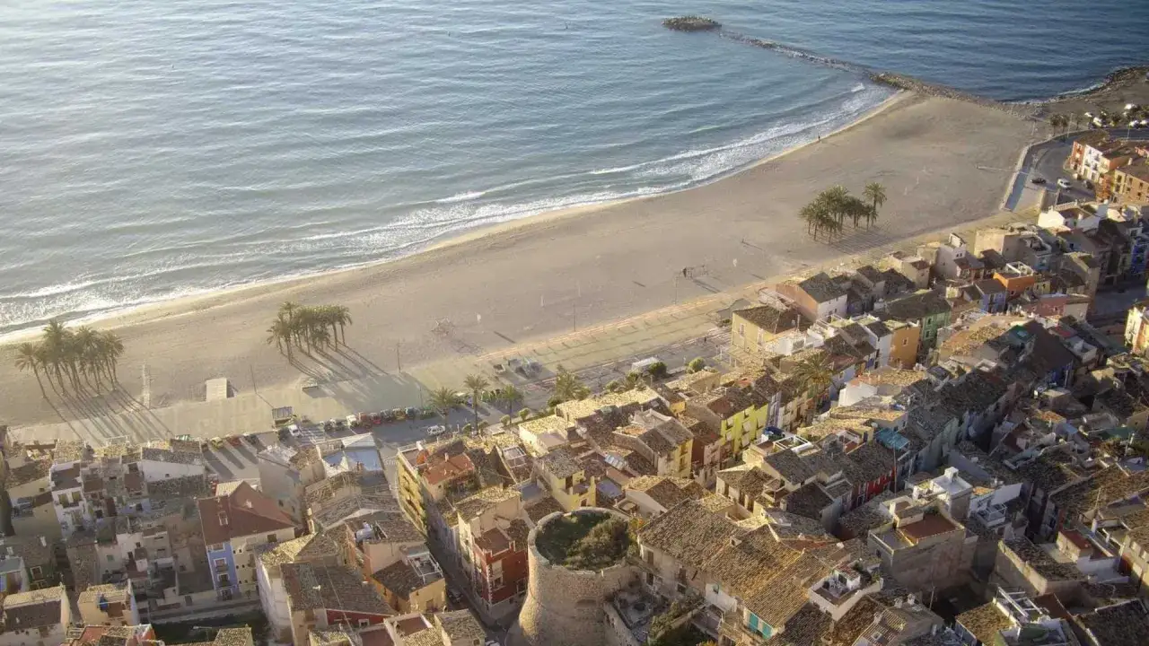 Vista a&eacute;rea de un pueblo costero con casas coloridas, una playa de arena y el mar. Se ve un antiguo torre&oacute;n, como si fuera un escenario para el maror fest.