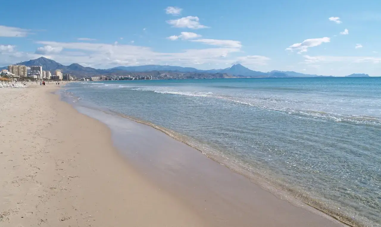 Arena dorada y mar azul en la playa Muchavista, con montañas y edificios al fondo bajo un cielo despejado.