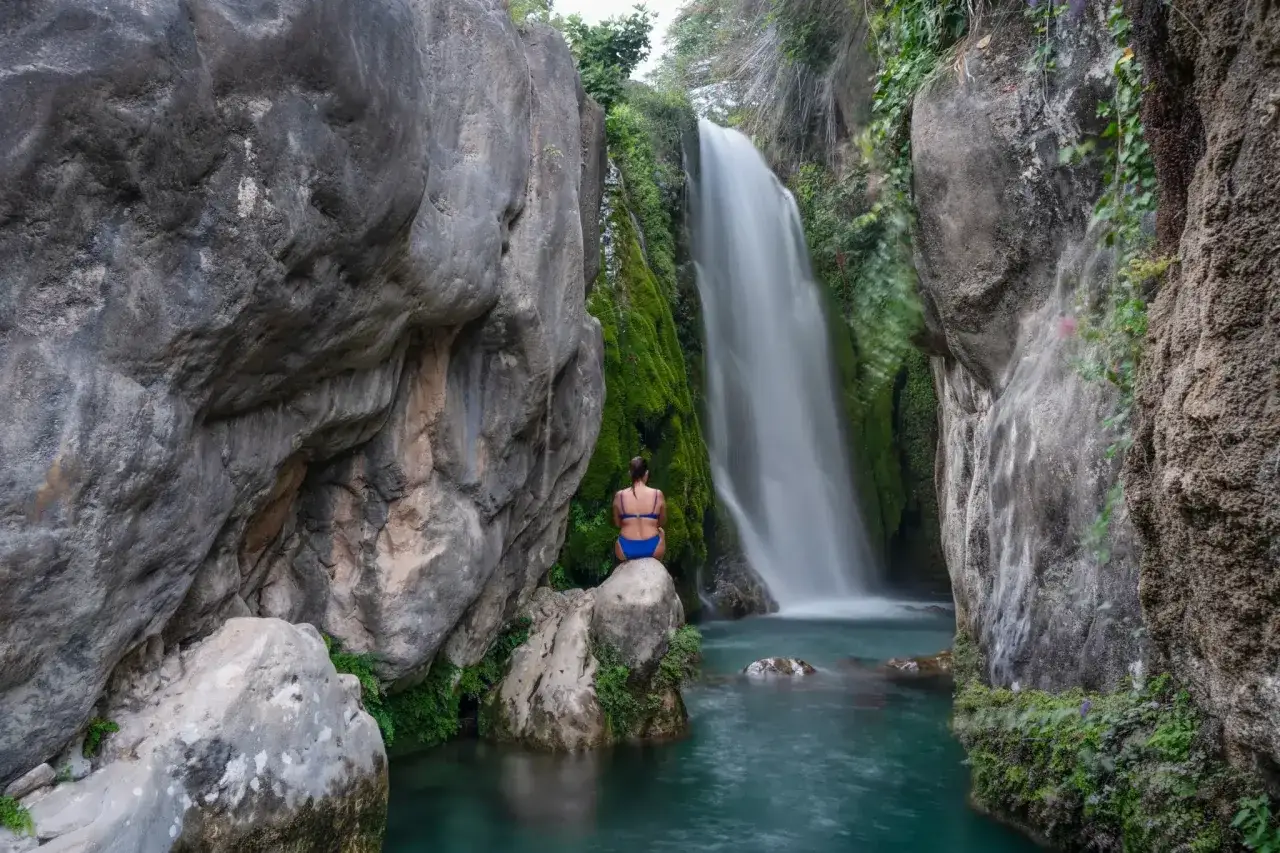 Una mujer sentada en una roca contempla la cascada de los fonts de Algar, rodeada de vegetación y rocas.