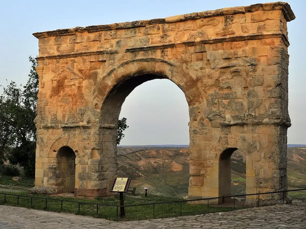 Imponente arco romano de piedra con dos arcos menores a los lados, sobre un paisaje de colinas verdes.