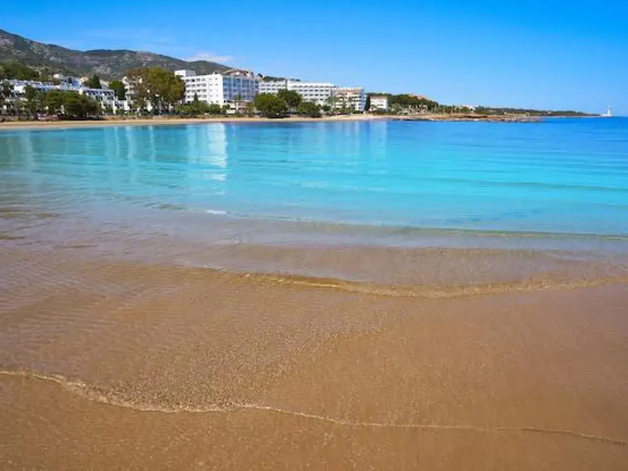 Aguas turquesas bañan la arena dorada de tres playas de Alcossebre, con hoteles y montañas al fondo bajo un cielo azul.