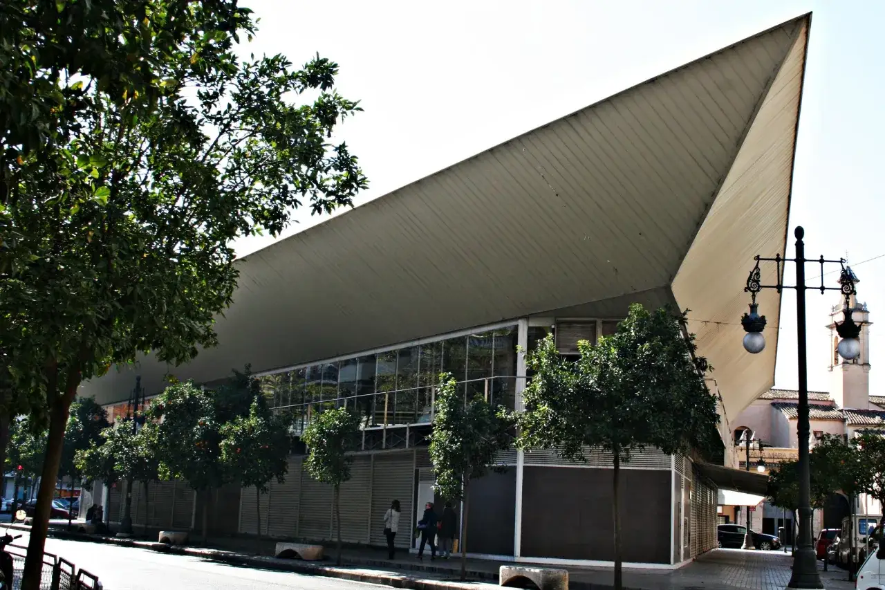 Edificio moderno con techo triangular en el mercado de Jesús. Árboles verdes bordean la calle y una farola ornamentada.