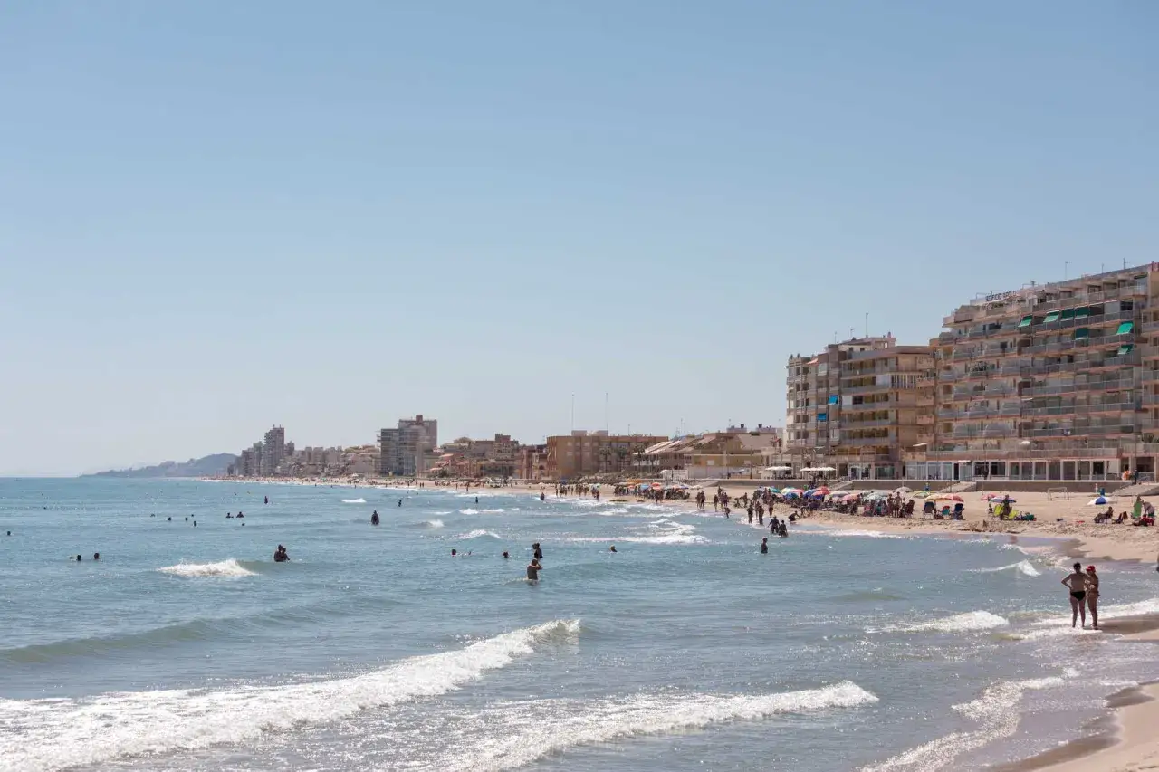 Gente disfrutando del sol y el mar en la playa del Perelló, con edificios residenciales al fondo.