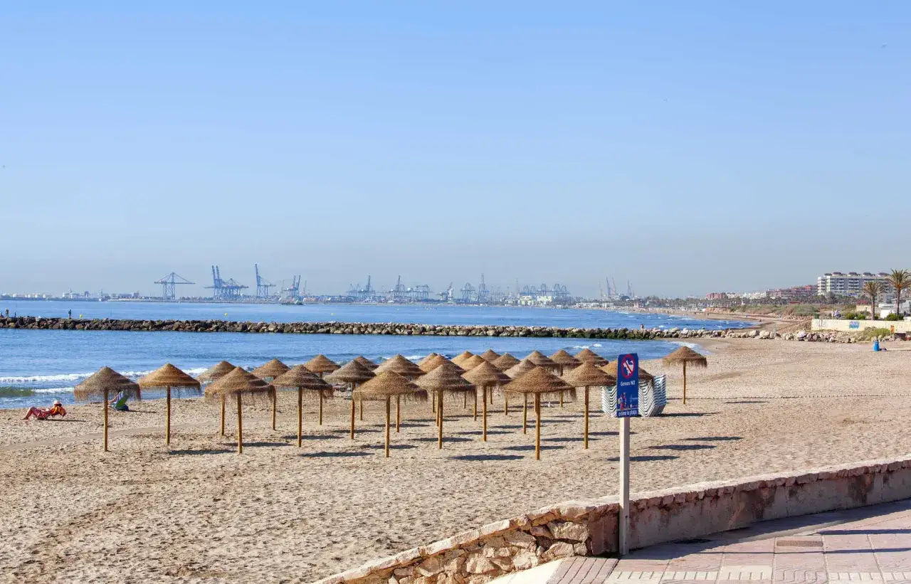 Sombrillas de paja en la playa port saplaya, con grúas de puerto y edificios al fondo.