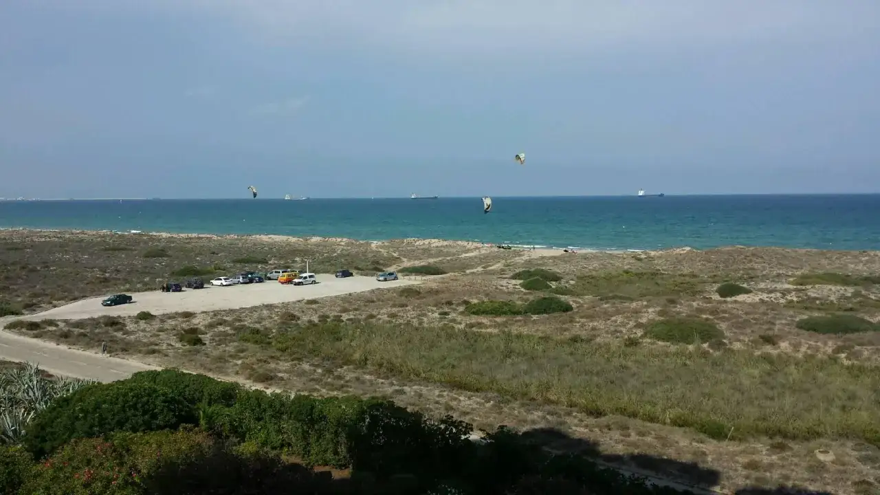 Dunas y mar azul en El Saler. Coches aparcados cerca de la playa, y kitesurfistas en el agua.