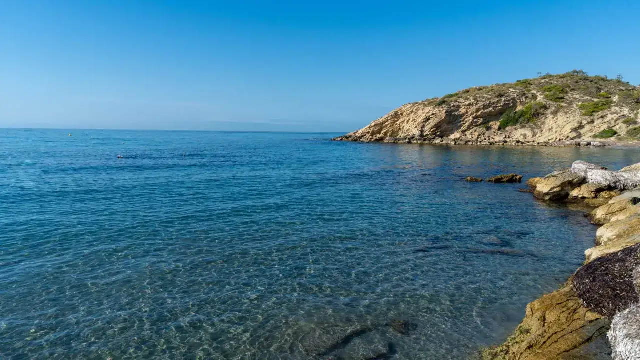 Aguas cristalinas en cala d'enmig, con una colina rocosa y vegetación al fondo. El mar azul se funde con el cielo despejado.