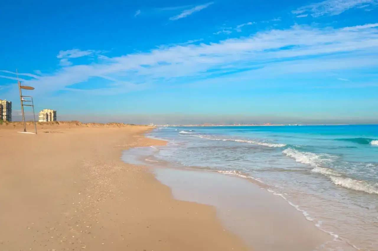 Amplia playa de arena dorada con el mar turquesa y edificios al fondo. ¡Podría ser la mejor playa de Valencia!