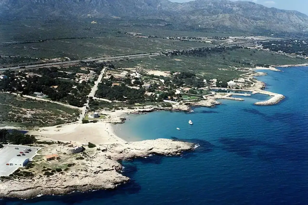 Vista aérea de la cala vidre, con su playa de arena dorada, aguas cristalinas y rocas escarpadas. Al fondo, montañas verdes y un pequeño puerto.