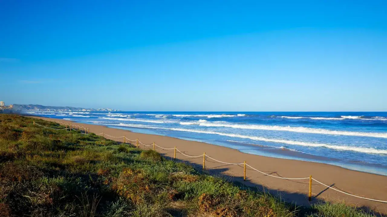 La playa de la goleta se extiende bajo un cielo azul intenso, con olas rompiendo suavemente en la arena dorada. Dunas cubiertas de vegetación bordean el camino.