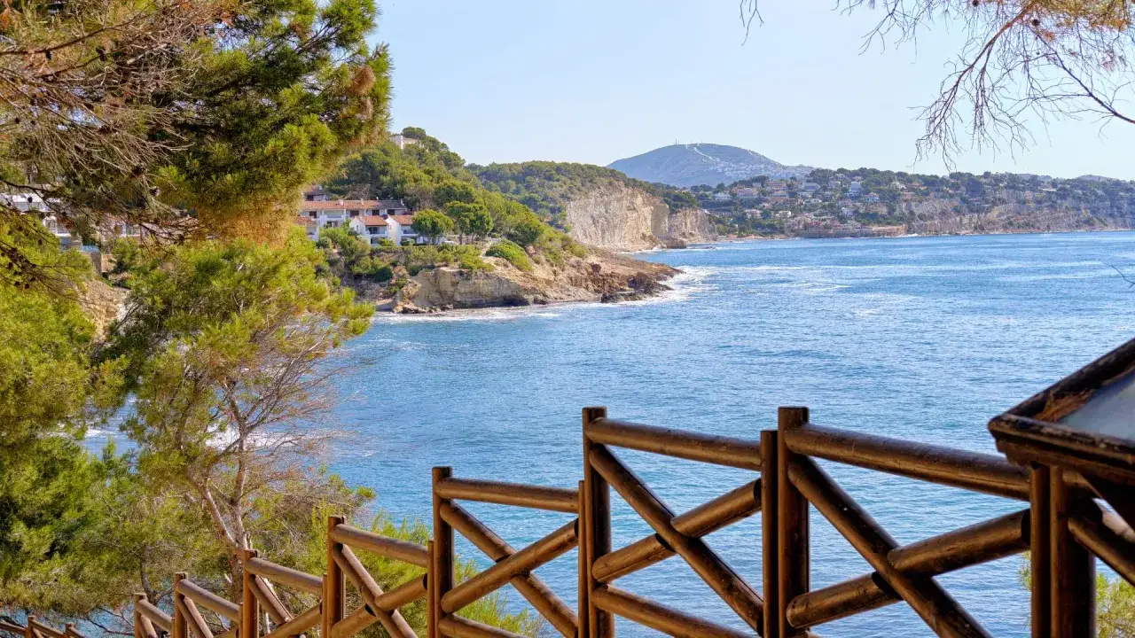Vista de la costa y el mar azul desde la cala Baladrar, con pinos y una valla de madera en primer plano.