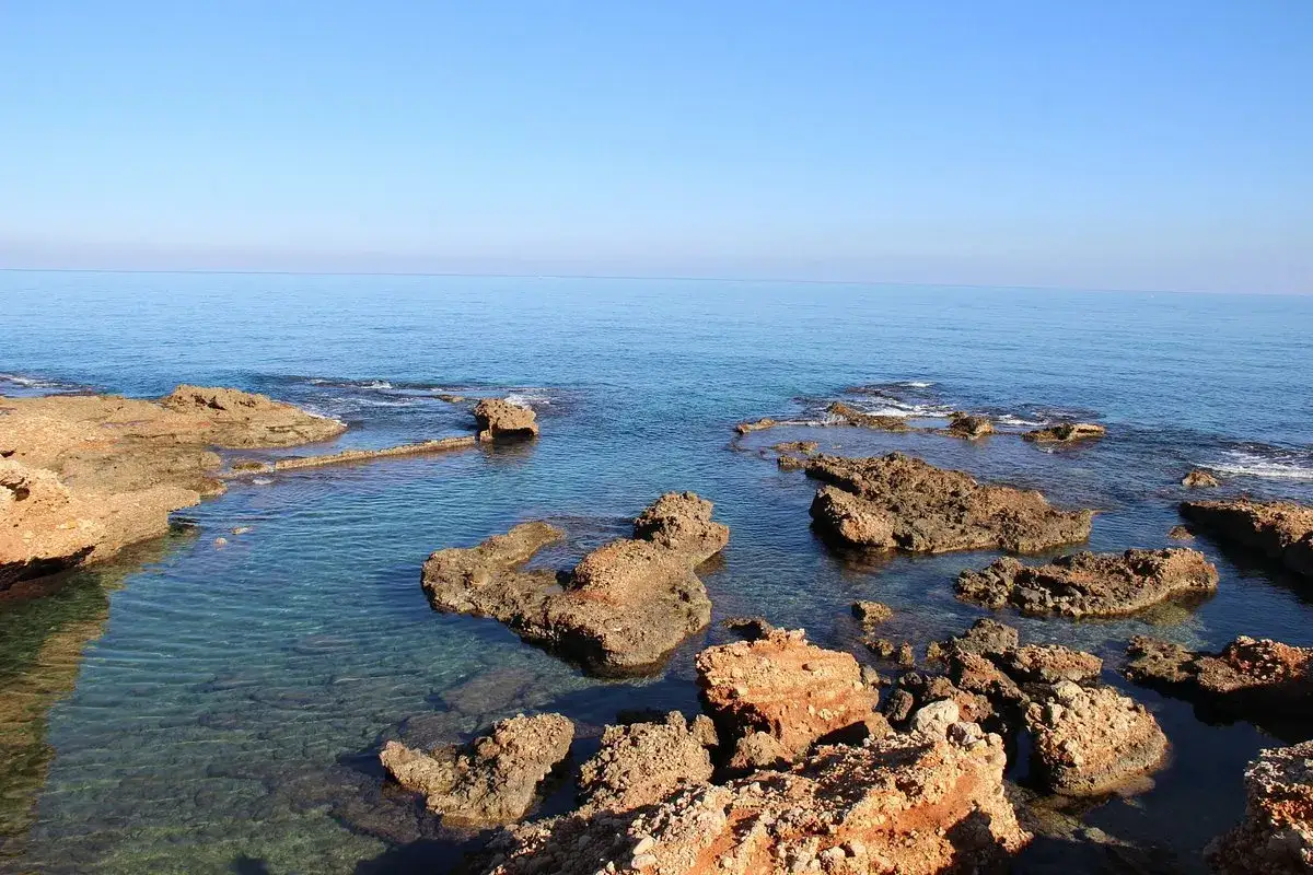 Aguas cristalinas y rocas rojizas en una de las mejores calas de Dénia. El mar Mediterráneo se extiende hasta el horizonte bajo un cielo azul.