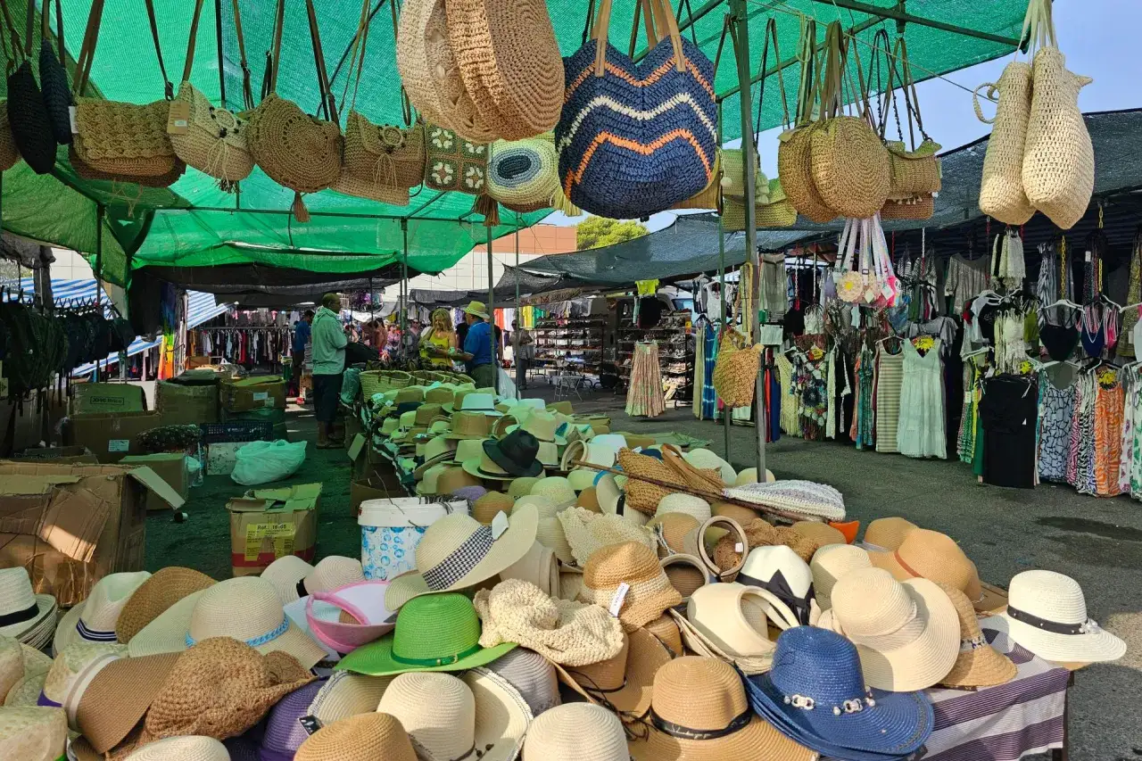 Sombreros y bolsos de mimbre cuelgan en un puesto del mercadillo Gran Alacant. Ropa y otros artículos se exhiben bajo toldos verdes.