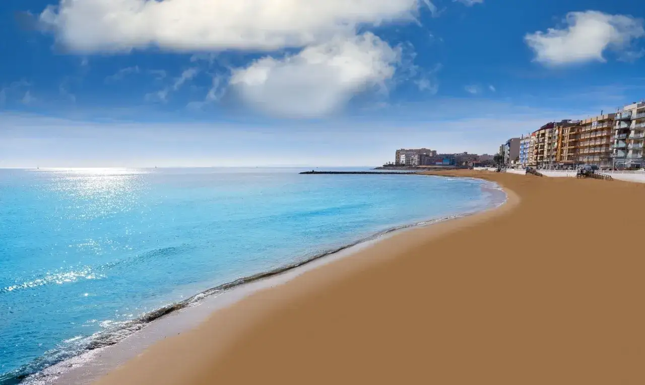 La playa de los locos se extiende dorada bajo un cielo azul con nubes algodonosas. El mar turquesa acaricia la arena, mientras edificios modernos se alinean en el horizonte.