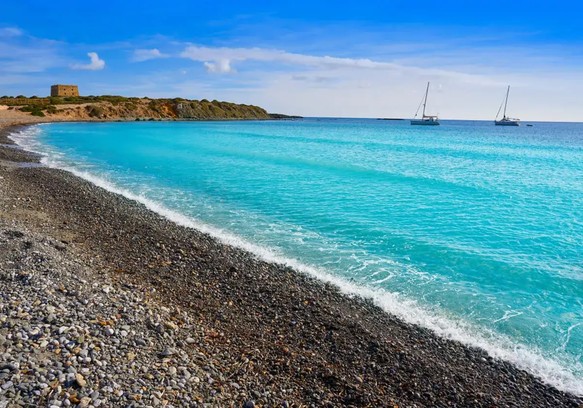 Playa de guijarros y aguas turquesas en la isla de Tabarca que ver. Dos veleros navegan cerca de la costa, con una torre defensiva en la colina.