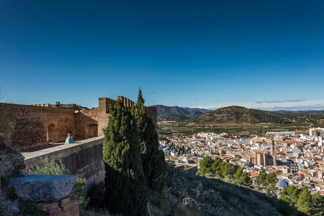 Vista panorámica del castillo de Sagunto con una persona contemplando el paisaje urbano y las montañas al fondo.