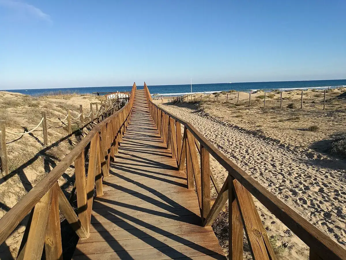 Pasarela de madera que conduce a la playa del campo, con dunas y el mar azul al fondo.
