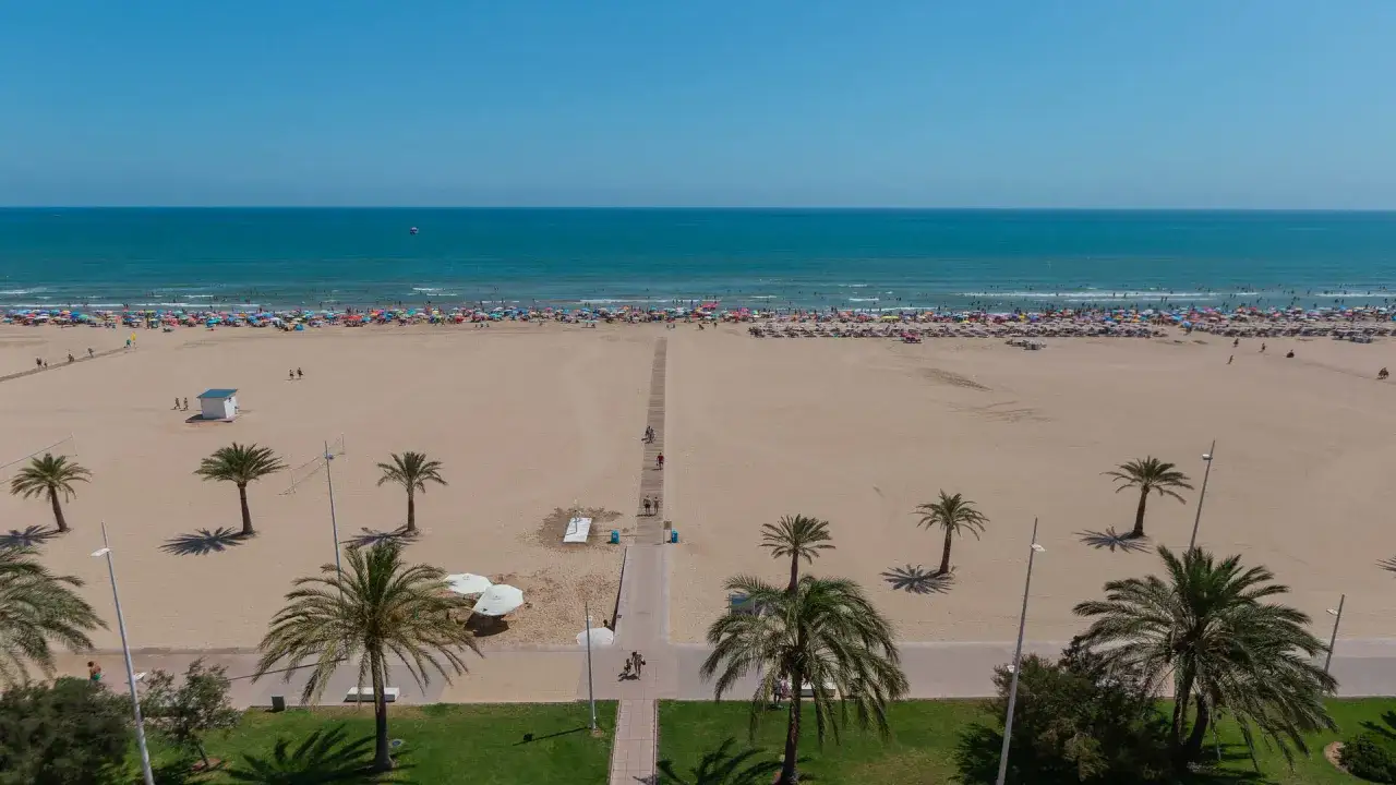 Amplia playa nord repleta de sombrillas y gente disfrutando del mar y la arena. Palmeras y un paseo marítimo completan la escena.