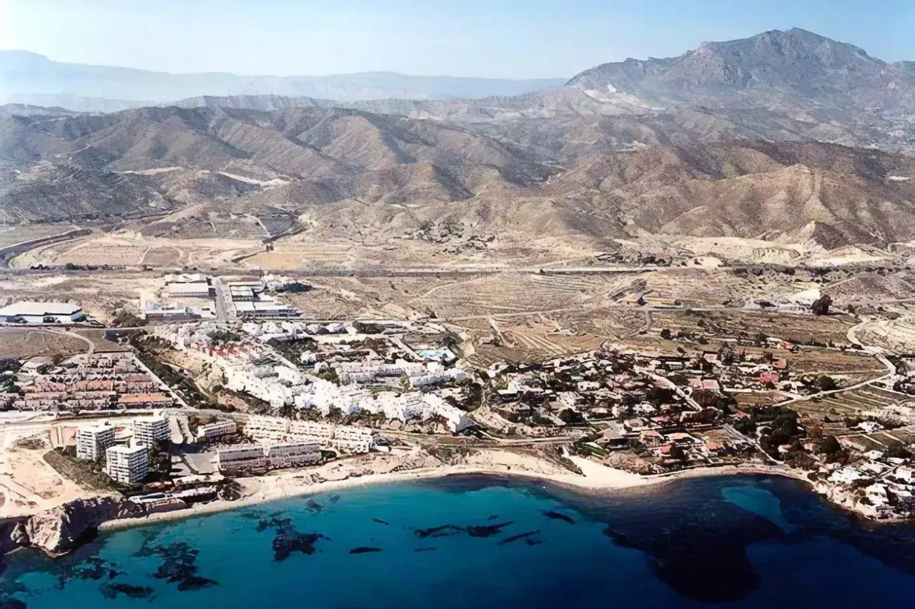 Vista aérea de la cala Amerador, con edificios blancos junto a la playa y montañas áridas al fondo.