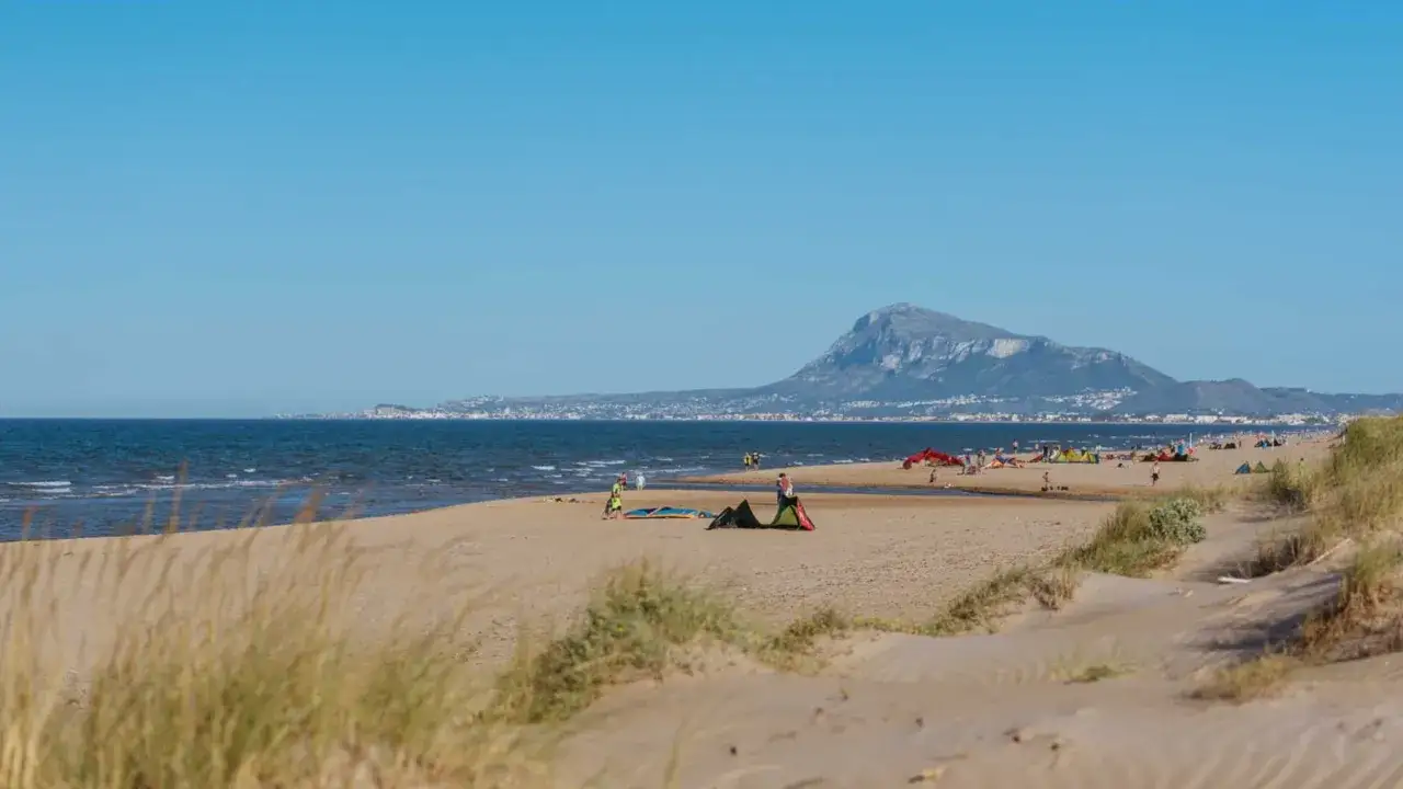 Amplia playa de arena con dunas y vegetación. El mar azul se extiende hasta el horizonte, donde se divisa una ciudad costera y una montaña. Varias personas disfrutan del día, algunas con equipos de kitesurf, en esta bega de mar.