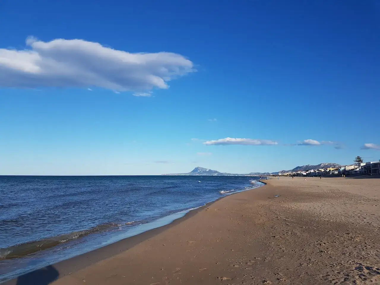 La playa Pau Pi, con arena dorada y mar azul, se extiende hacia un horizonte montañoso bajo un cielo despejado con nubes algodonosas.