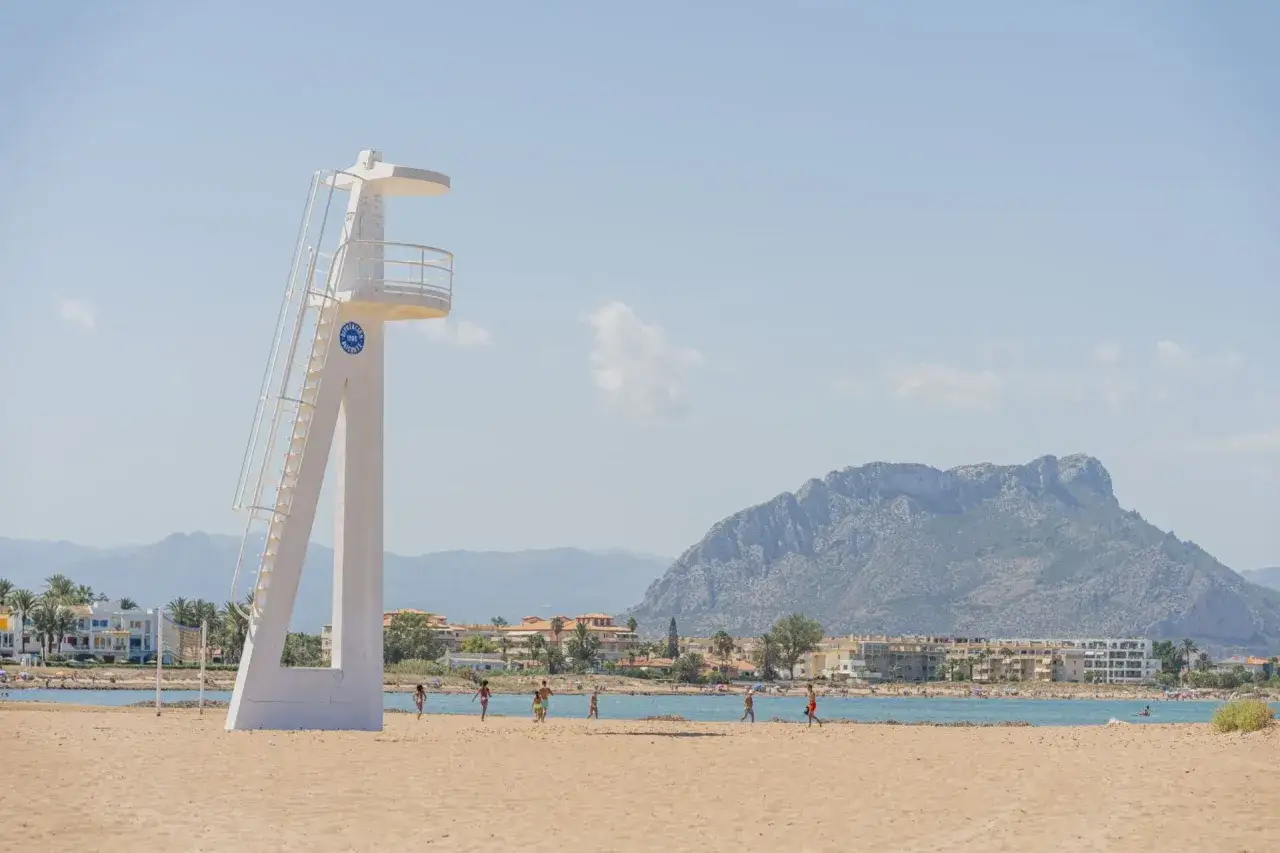 Torre de vigilancia en la playa Els Molins, con niños jugando en la arena y un imponente monte al fondo.