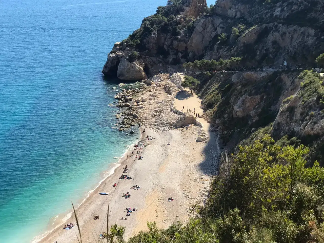 Playa de guijarros con aguas turquesas, acantilados y un camino que lleva a la Cova dels Arcs. Gente disfrutando del sol.