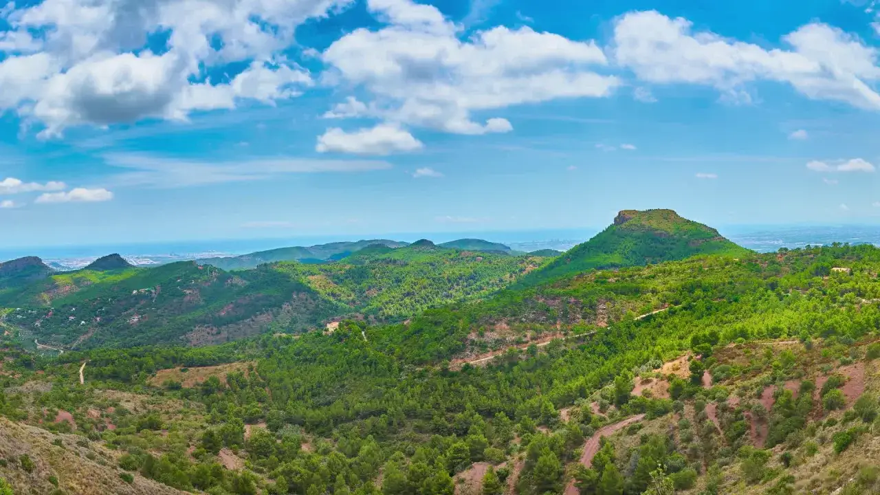Paisaje montañoso verde con el mar al fondo, ideal para camping en la Sierra Calderona. Nubes blancas en cielo azul.