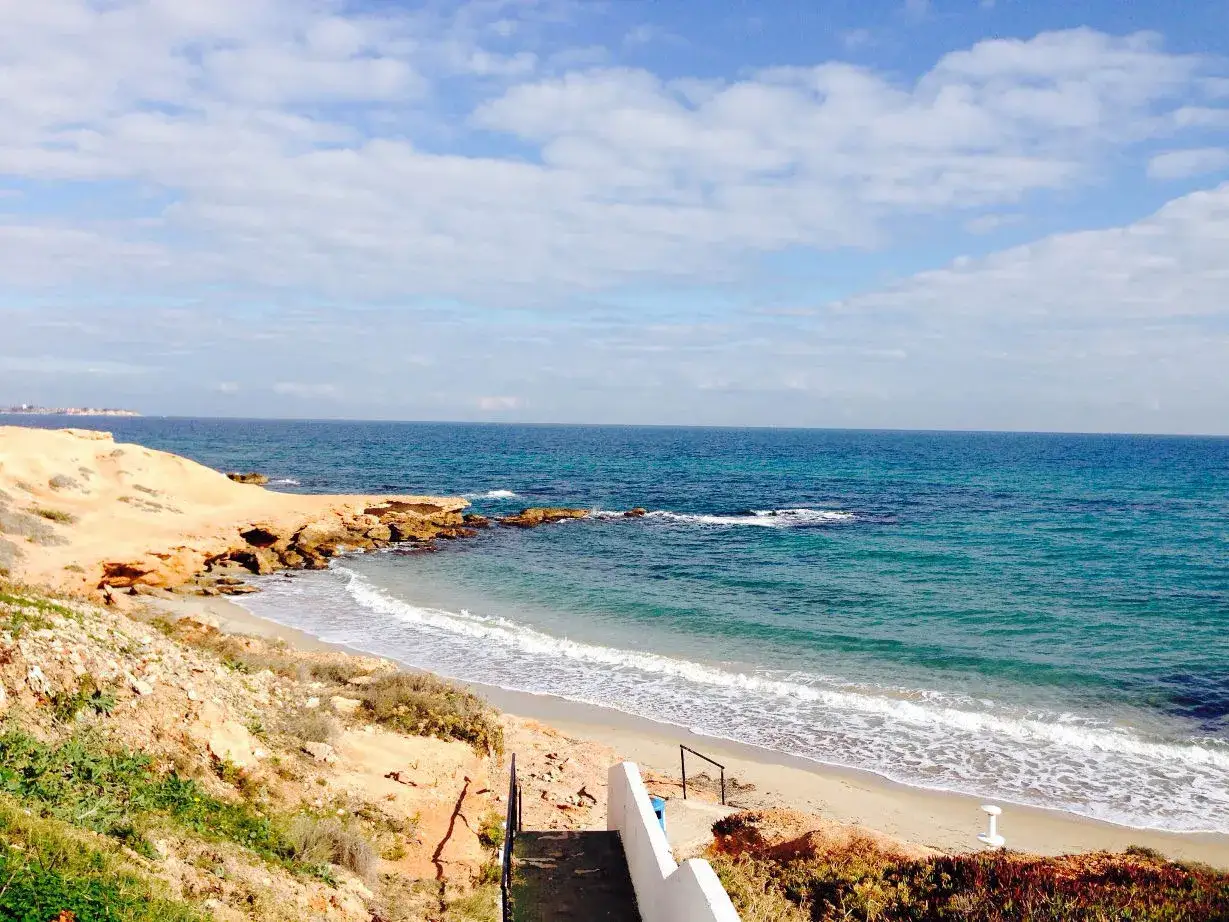 Escaleras descienden a la playa rocamar, con arena dorada, rocas y el mar azul bajo un cielo nublado.