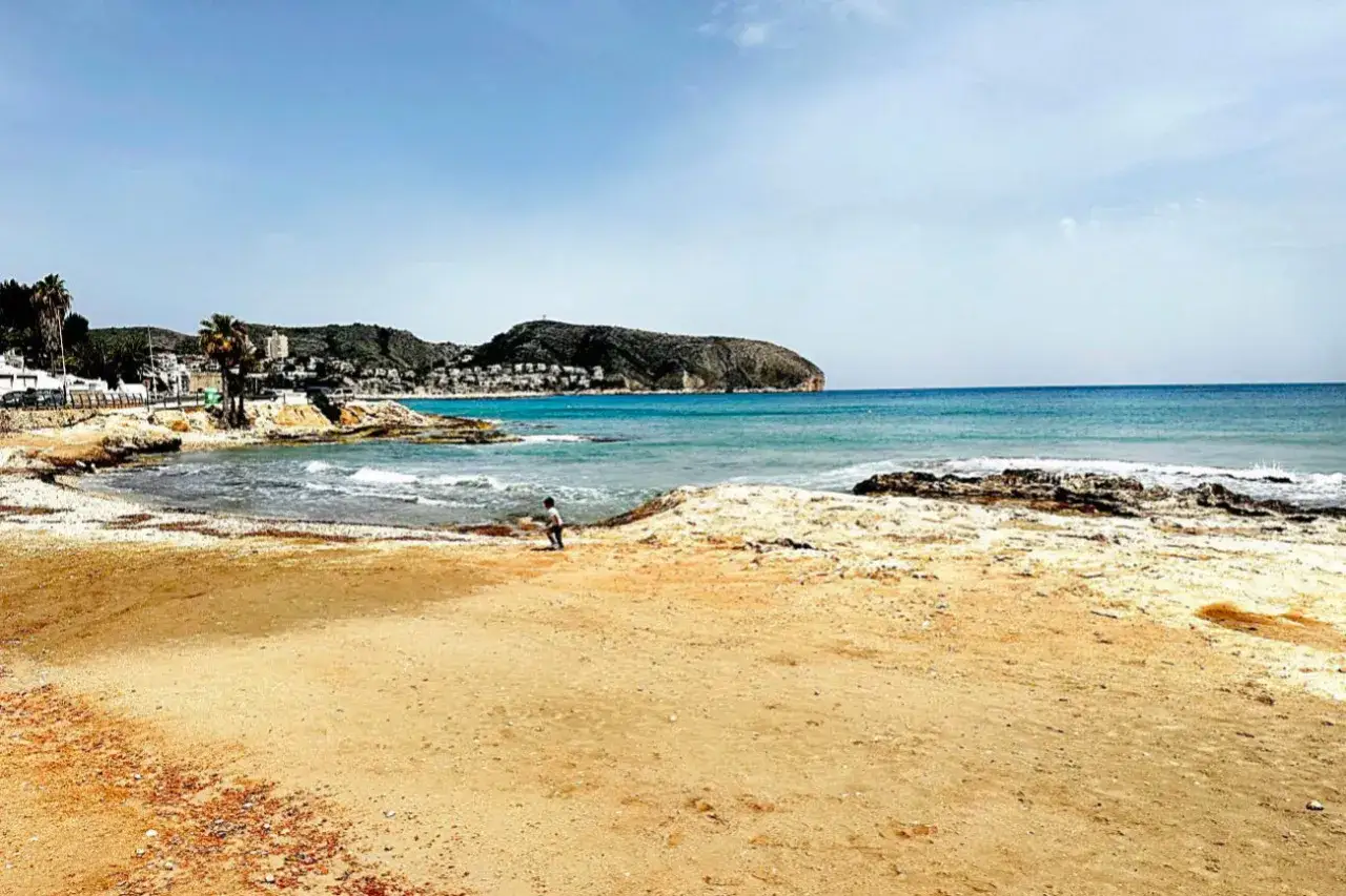 Ni&ntilde;o juega en la arena dorada de cala l'andrag&oacute;, con el mar azul y monta&ntilde;as al fondo.