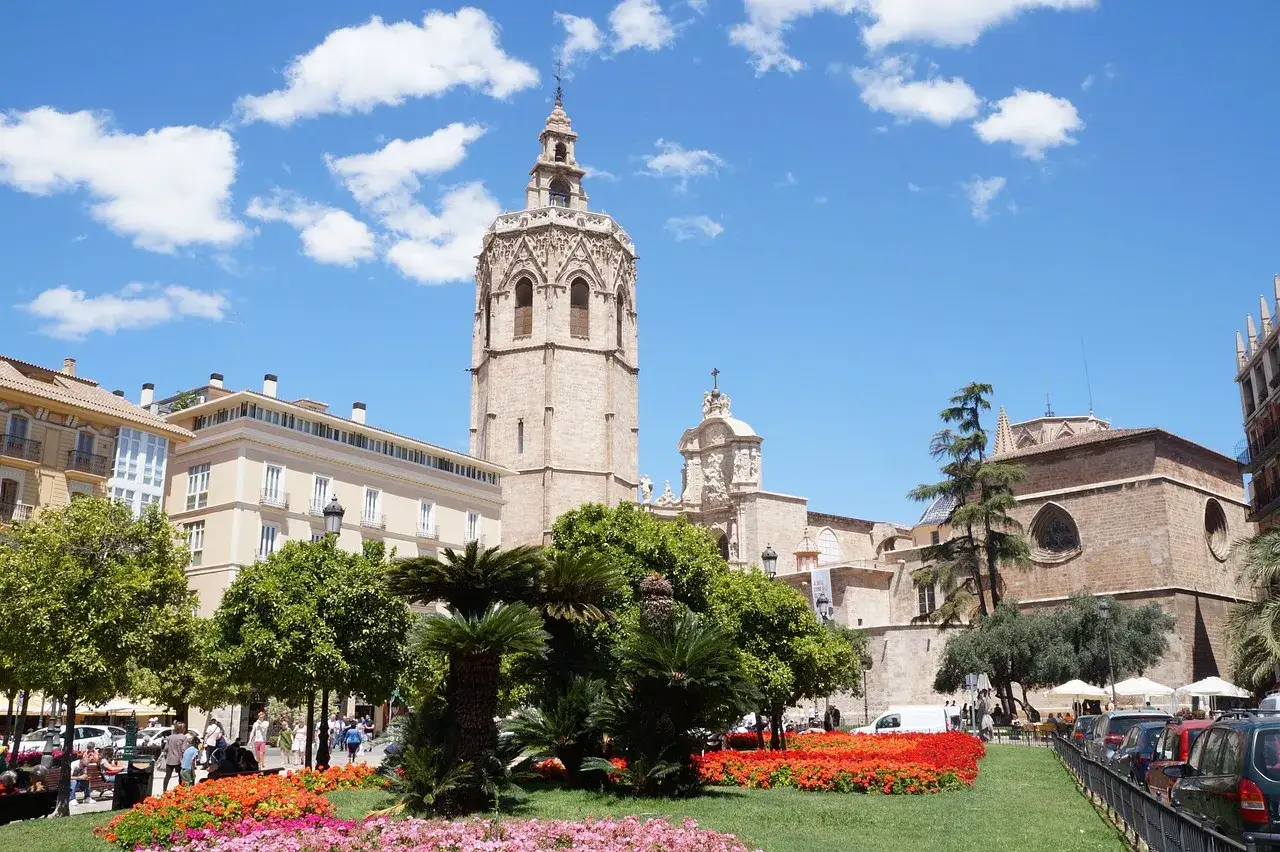 El Miguelete, campanario de la Catedral de Valencia, se alza majestuoso sobre un jardín florido y edificios históricos bajo un cielo azul con nubes.