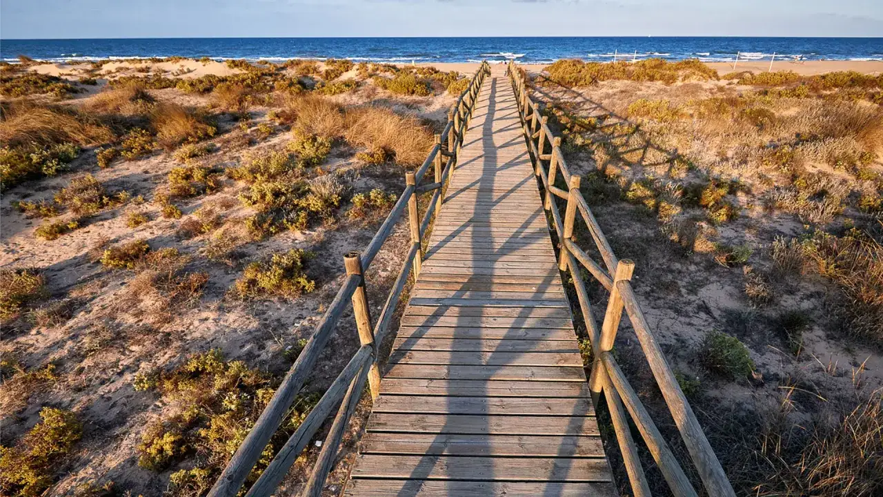 Pasarela de madera que conduce a la playa de l'Ahuir, rodeada de dunas y vegetación costera. El mar azul se extiende hasta el horizonte.