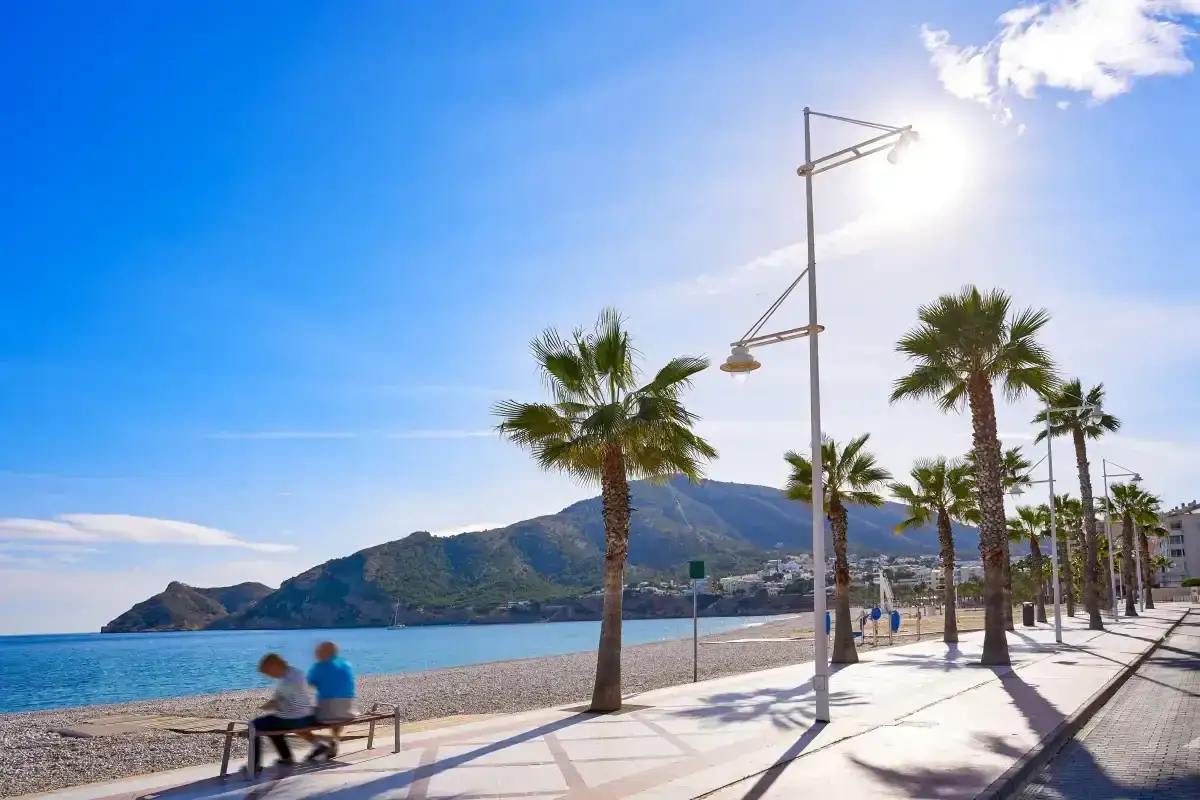 Pareja sentada en un banco en la playa cap blanc, con palmeras y montañas al fondo bajo un cielo azul brillante.
