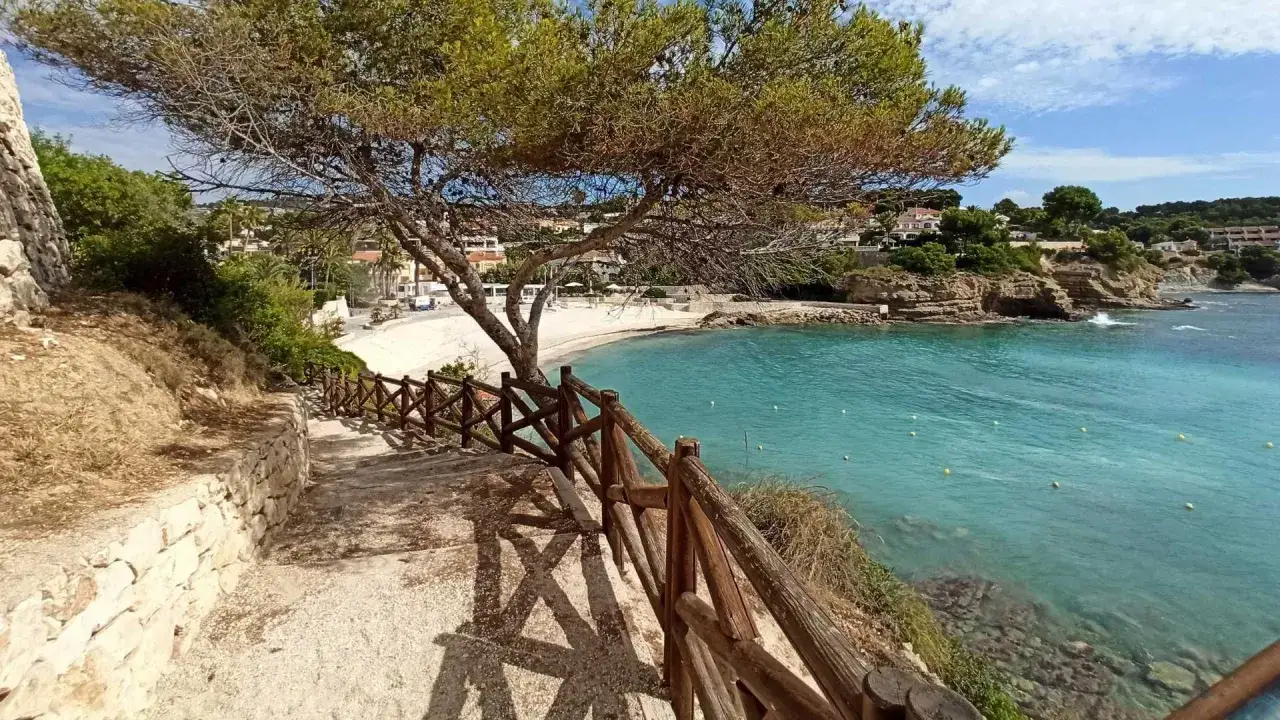 Escaleras de madera bajan hacia la cala Pinets, con aguas turquesas y una playa de arena blanca.