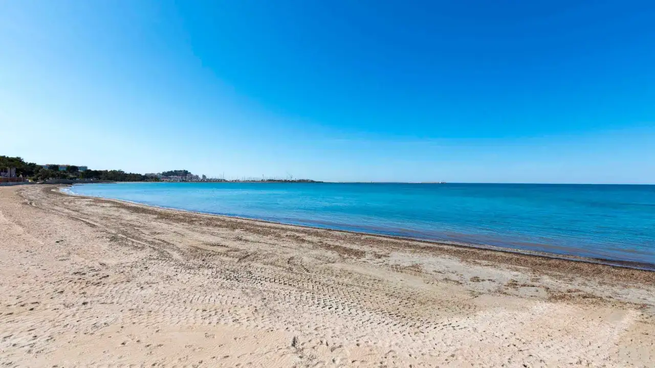 Arena dorada y mar azul turquesa en la playa de la Marineta Cassiana, con el horizonte de la ciudad al fondo.