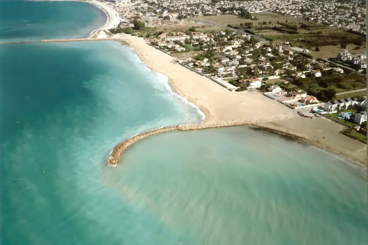 Vista aérea de la playa Deveses con aguas turquesas y rompeolas de piedra. Casas blancas y vegetación bordean la costa.