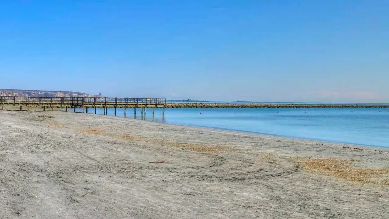Paseo de madera sobre la arena gris de la playa La Gola, Santa Pola, con el mar azul y la ciudad al fondo.