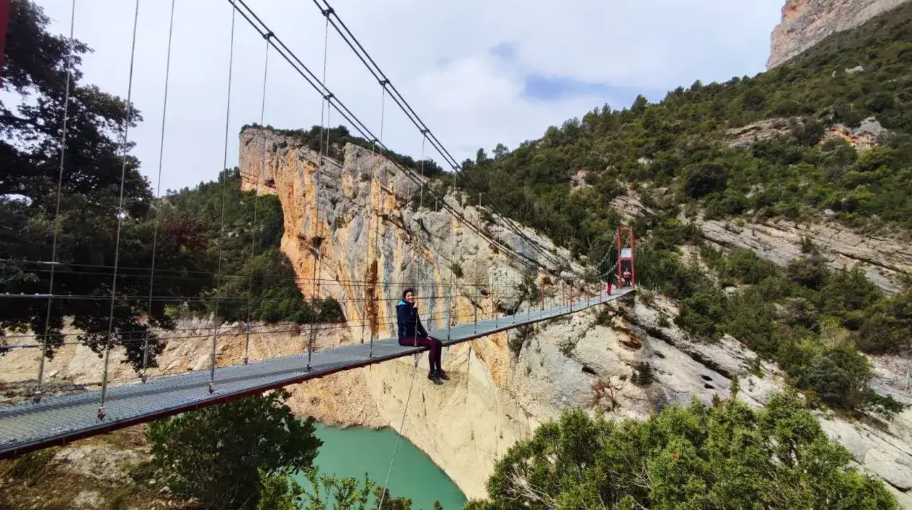 Persona sentada en el puente colgante del desfiladero de Mont-rebei, con vistas a un cañón y un embalse de agua turquesa.