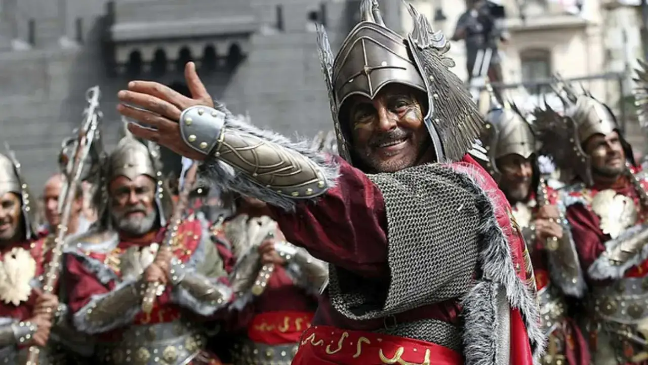 Un hombre sonriente con armadura y casco alado saluda, liderando un desfile de moros y cristianos.