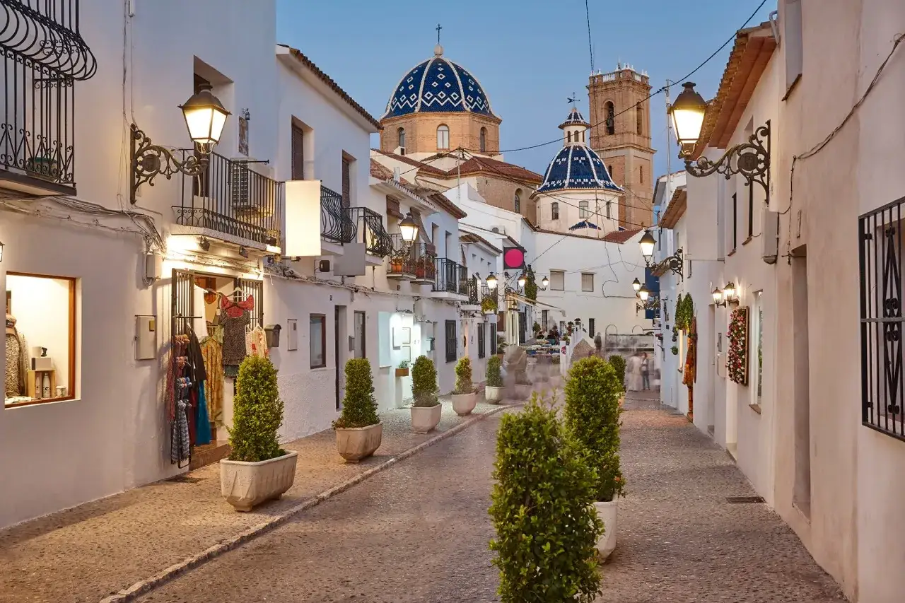 Calle empedrada en la altea vieille ville, con casas blancas, farolas y cúpulas azules al fondo.