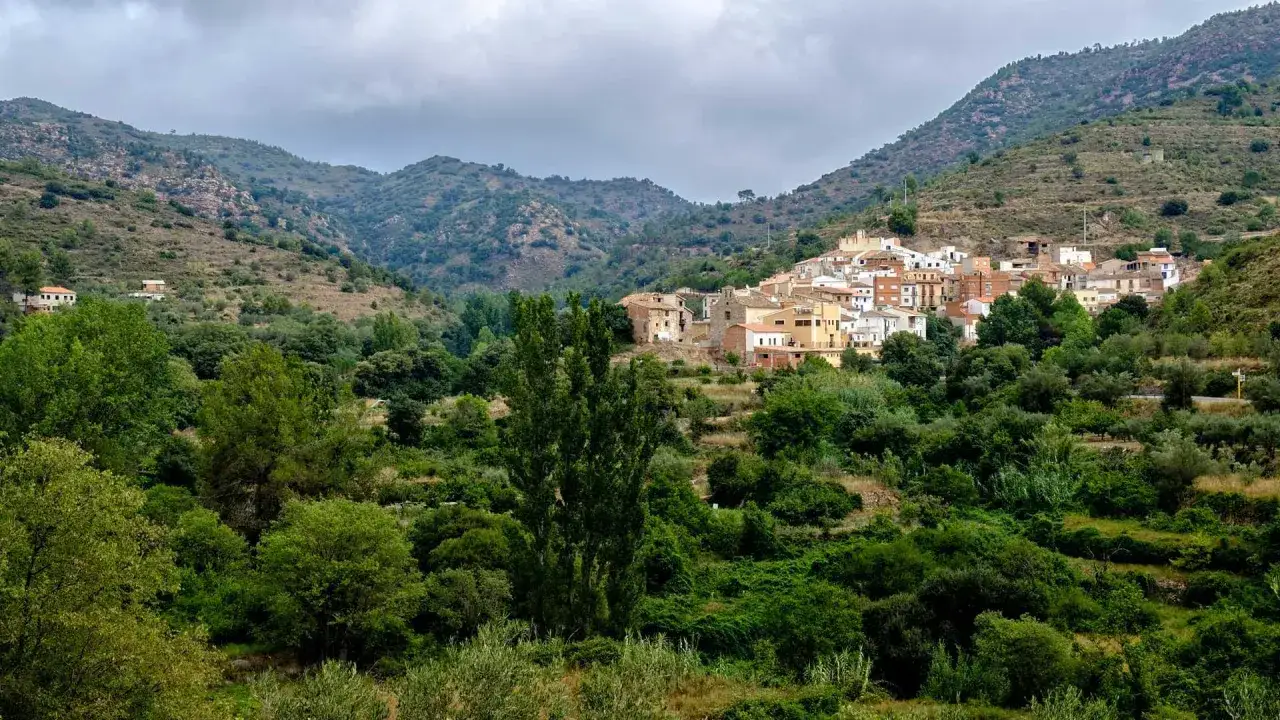 Pueblo blanco anidado en la verde sierra de Espadán, rodeado de vegetación exuberante y colinas onduladas bajo un cielo nublado.