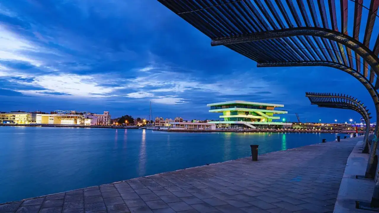 El puerto de Valencia al anochecer, con el edificio Veles e Vents iluminado y el reflejo de las luces en el agua.