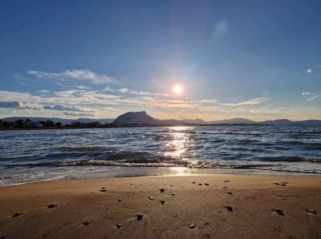 Atardecer en la playa de arena de Dénia. El sol brilla sobre el mar Mediterráneo, reflejándose en las olas. Montañas y edificios se vislumbran en el horizonte.
