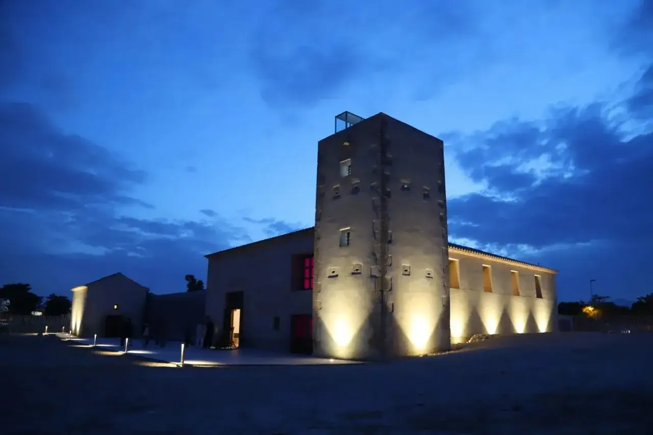 La torre Ansaldo, iluminada al anochecer, se alza junto a edificios bajos bajo un cielo azul oscuro con nubes.