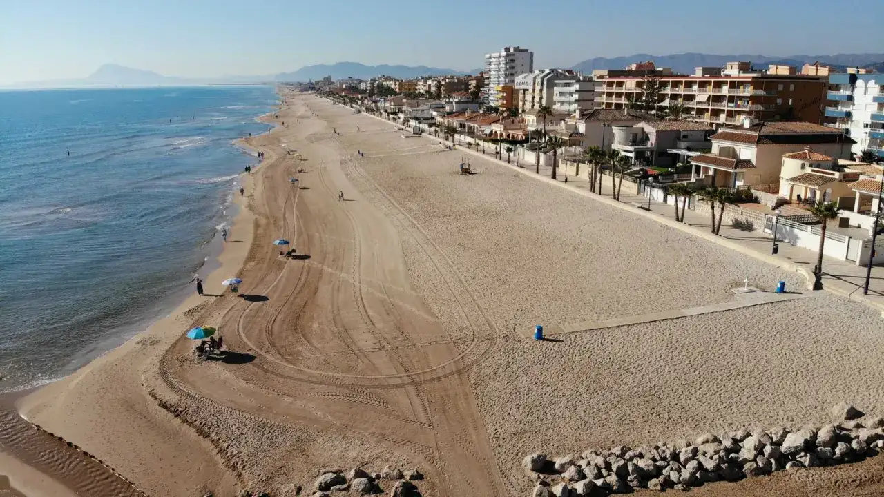 Amplia playa de Miramar con arena dorada, mar azul y edificios turísticos al fondo. Pocas personas disfrutan del sol bajo sombrillas.