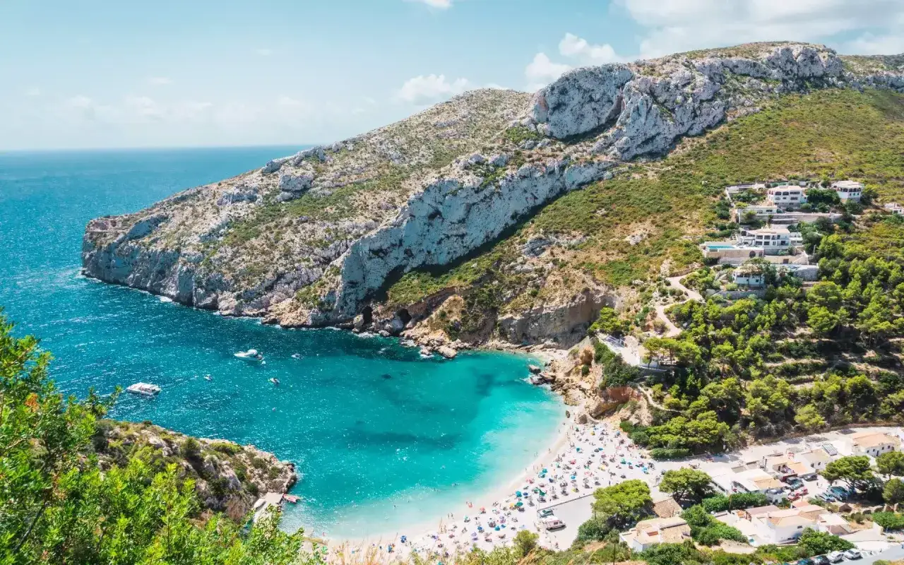 Bahía de aguas turquesas con una playa repleta de sombrillas y gente. Montañas escarpadas y casas blancas salpican la costa, un paraíso de calas alicante.