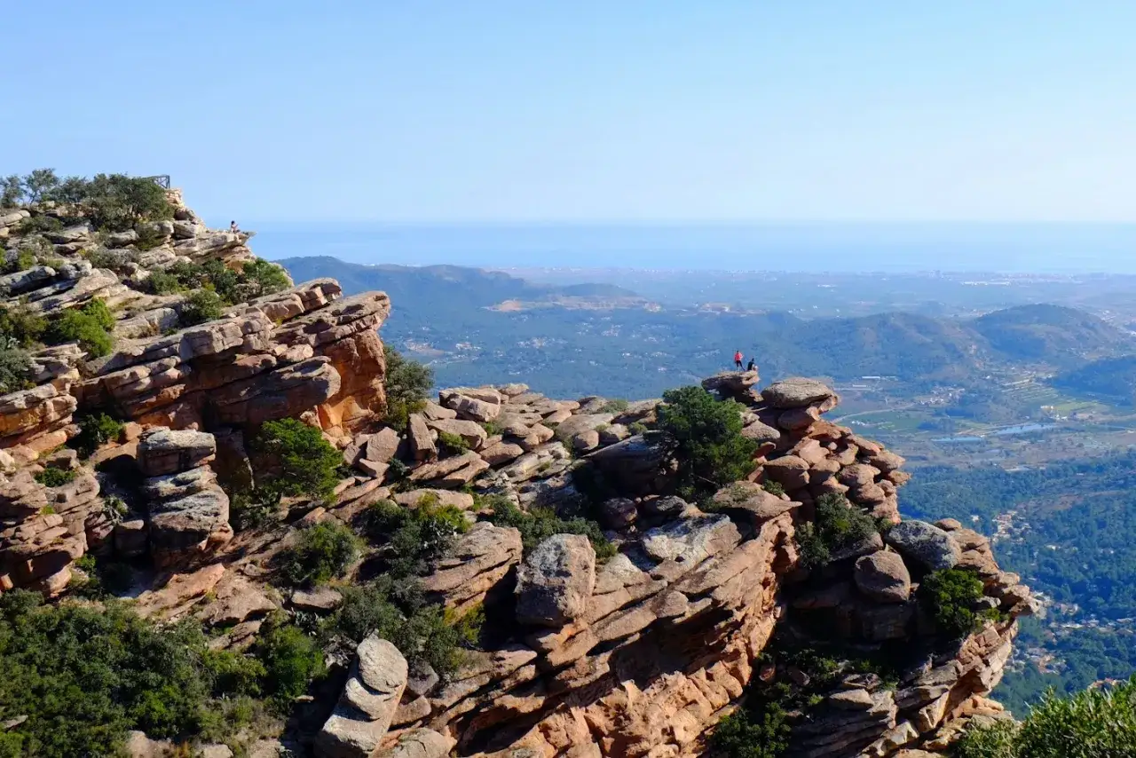Dos excursionistas disfrutan de la vista desde la cima rocosa de el garbi, con el mar azul al fondo.